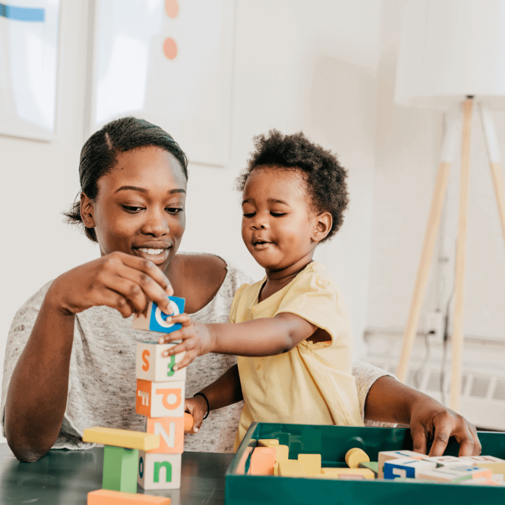 A mother and toddler play with colorful alphabet blocks, stacking them on a table in a bright room, conveying a sense of learning and possible early intervention.