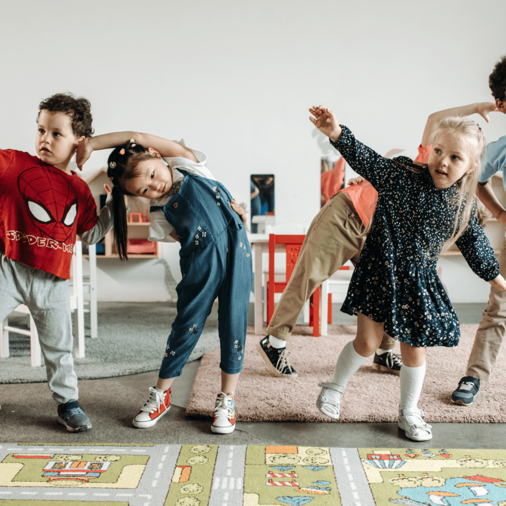 Children in colorful clothing playfully stretch in a classroom, exhibiting joy and energy. The room is bright, with rugs and small furniture. Early diagnosis in ASD.