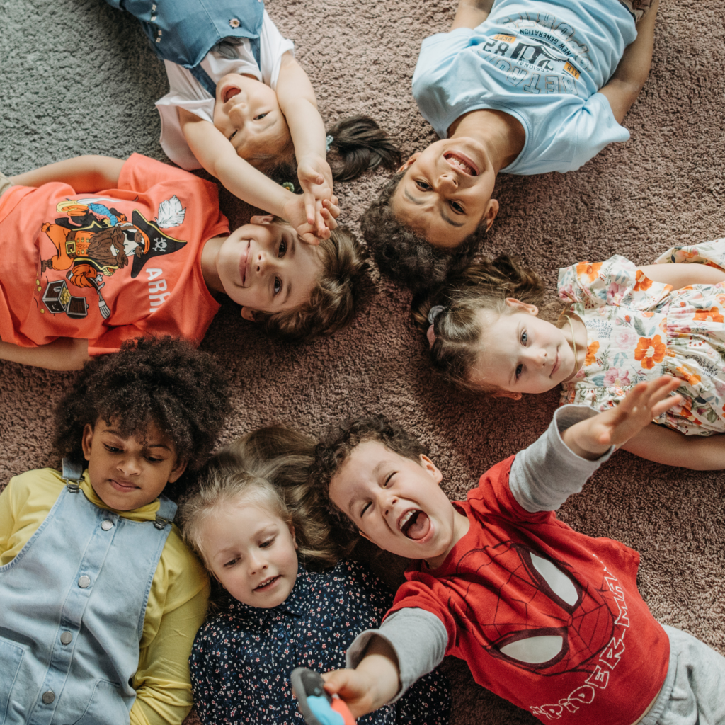 A group of seven children, lying on a carpet in a circular formation, smiling and laughing. Their heads meet at the center, creating a joyful scene. Shows how ABA can be useful beyond autism.