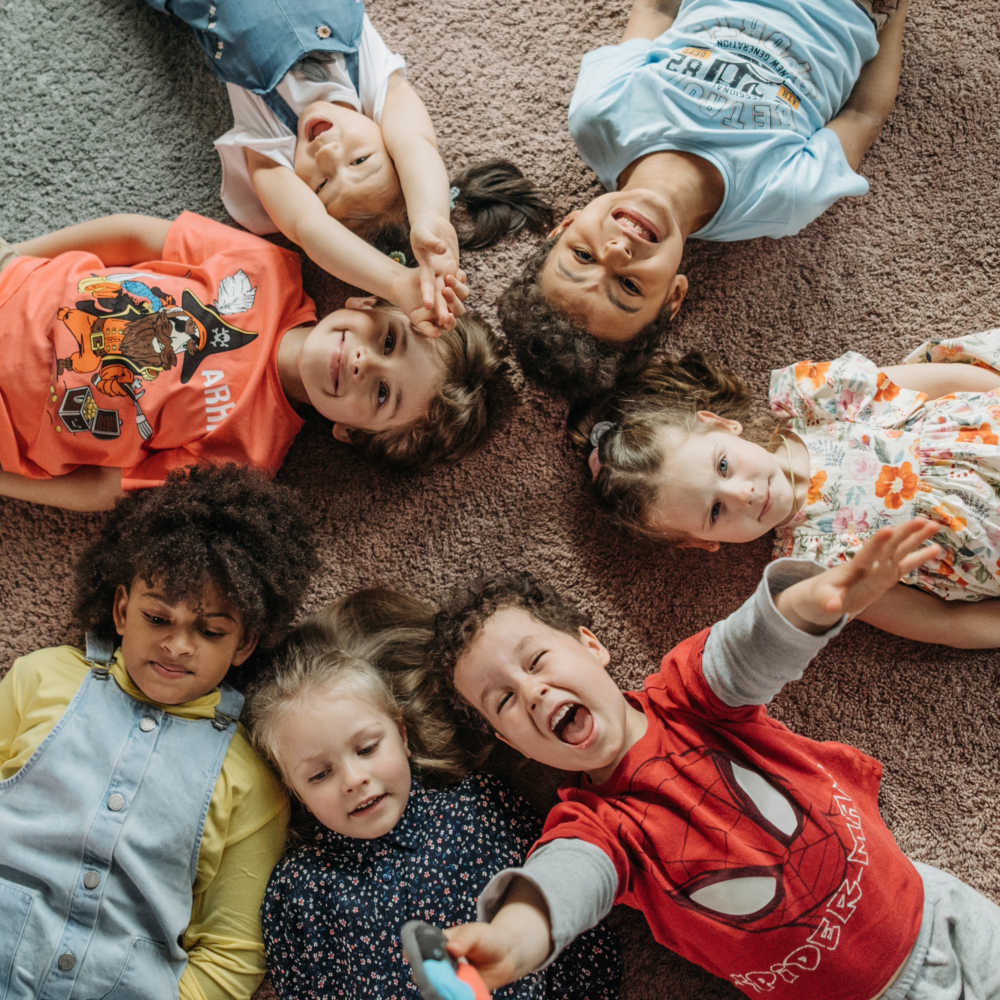 A group of seven children, lying on a carpet in a circular formation, smiling and laughing. Their heads meet at the center, creating a joyful scene. Shows how ABA can be useful beyond autism.