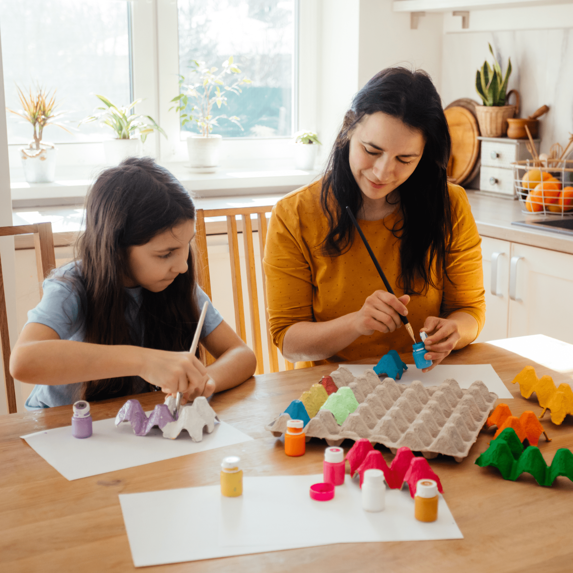 A woman and a child are painting egg cartons with bright colors at a wooden table. Sunlight streams through a window, creating a warm, creative atmosphere. One of many 155 activities for when you're stuck at home.