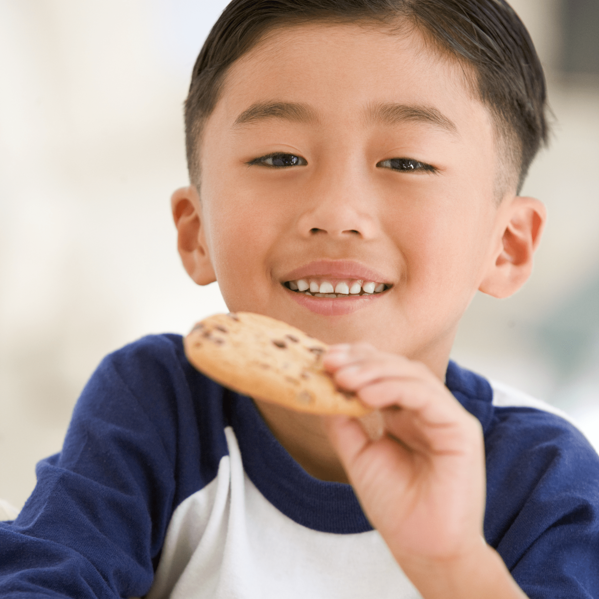 Young boy smiling and holding a chocolate chip cookie. He wears a blue and white shirt and appears happy and content, suggesting an enjoyable moment. Possible that mand training helped the child get the cookie.