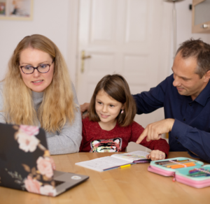 A family sits at a wooden table. A woman and a man, on either side of a young girl in a red sweater, engage with a floral laptop. The scene feels warm and focused.