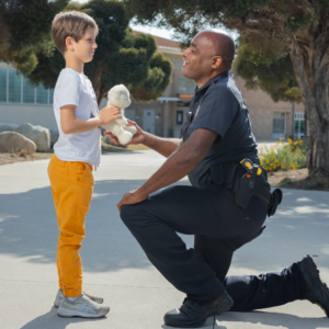 A police officer kneels, smiling warmly at a young boy, offering a stuffed animal. They are outdoors near a school building. Friendly, caring exchange.