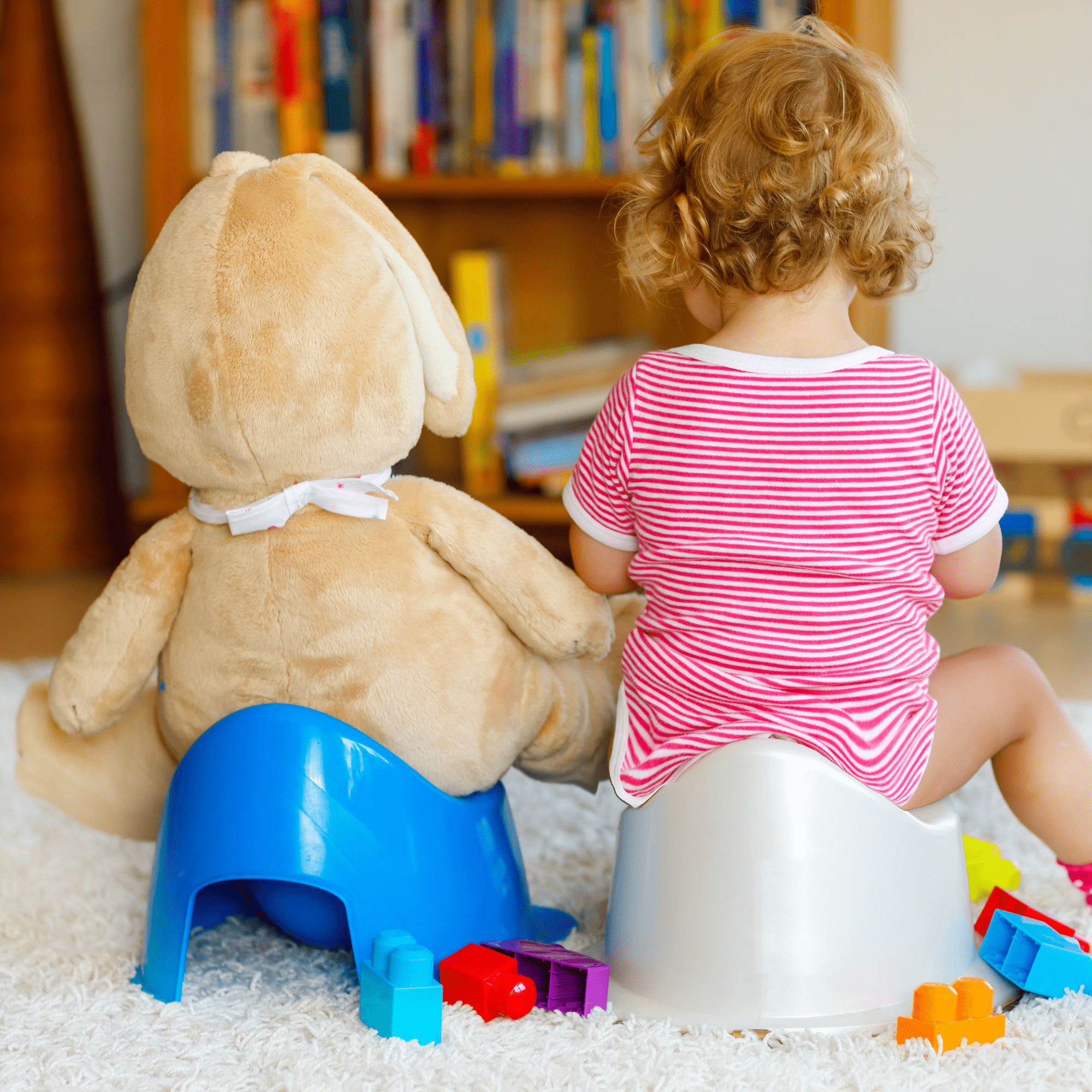 A toddler and plush bunny sit on training toilets, facing away, in a cozy room with books and toys scattered around. The tone is playful and warm.