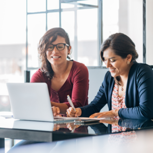 Two women sit at a table with a laptop. It looks like one is giving the other feedback. One writes while smiling; the other, wearing glasses, looks on. Bright, modern office setting.