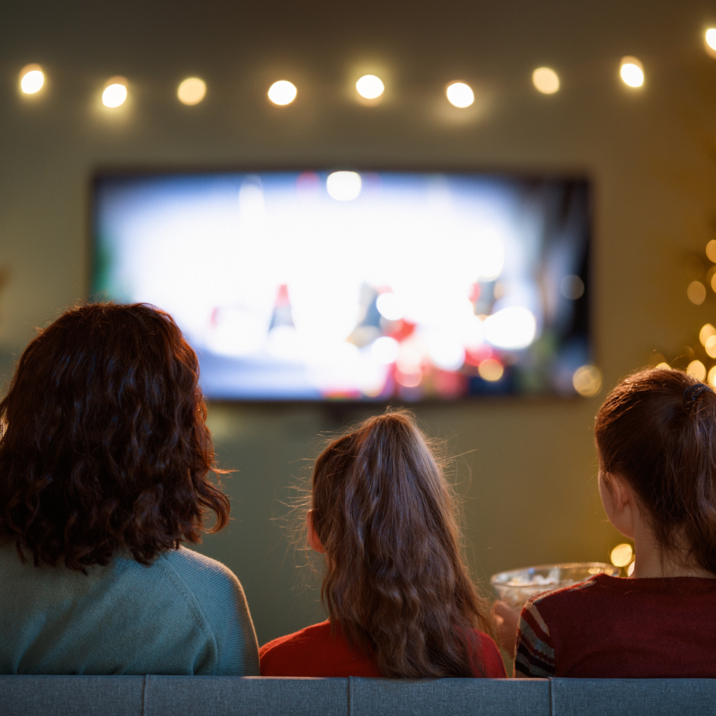 A family of three sits on a couch watching TV, backs facing the viewer. Warm lighting and a blurry screen create a cozy, festive atmosphere.