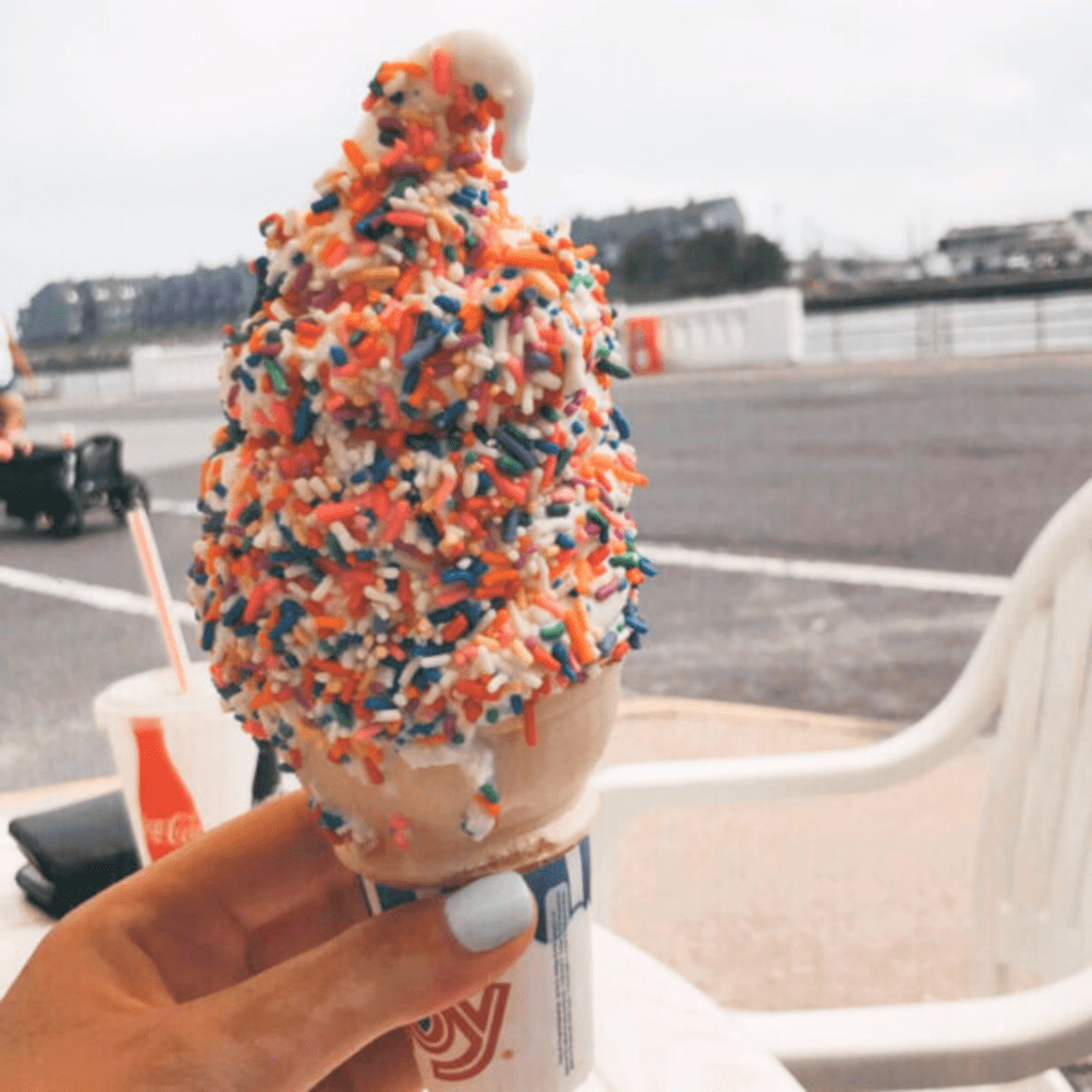 A hand holds a soft-serve ice cream cone covered in colorful sprinkles. The setting is outdoors with a road and distant buildings in the background.