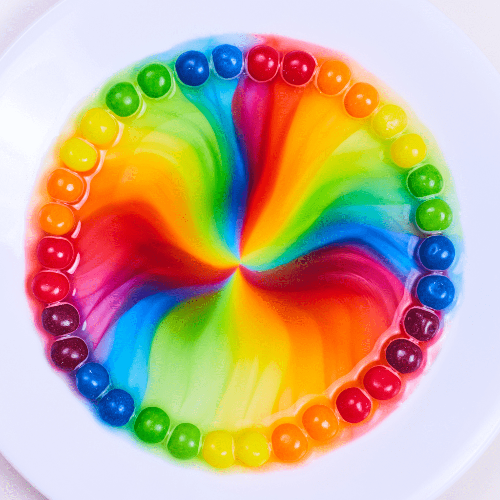 A white plate with colorful candies forming a circle along the edge. Inside, vibrant rainbow colors blend inwards, creating a swirling pattern.