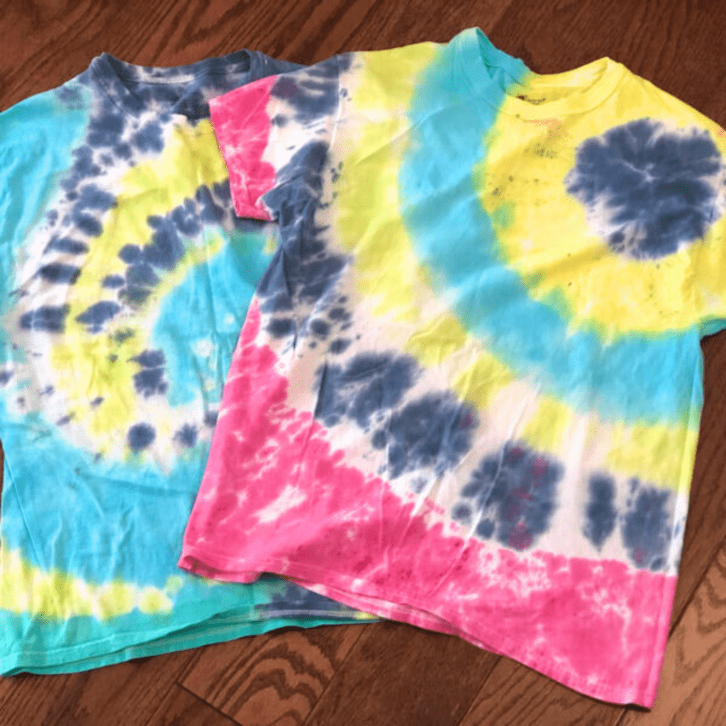 Two vibrant tie-dye shirts lie on a wooden floor. One features blue and yellow swirls, the other has spirals of pink, yellow, and blue. Bright and playful.