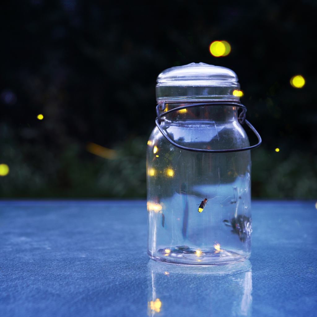 A glass jar on a reflective surface holds glowing fireflies, with a few flying outside. The dark, blurred background creates a magical atmosphere.