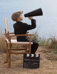 Young boy in director's chair by a lake, wearing a suit and cap, holds a megaphone. Clapperboard on ground; scene suggests imaginative play.