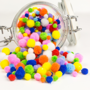 A glass jar tipped over with colorful pom-poms spilling out. The pom-poms are vibrant in various sizes, creating a playful, cheerful scene. building kindness and conversation