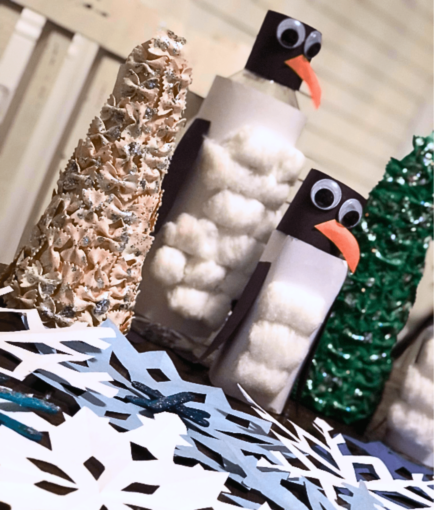 Festive table with handmade crafts: two penguins made of bottles and cotton, surrounded by paper snowflakes and pasta trees, conveying a winter vibe.