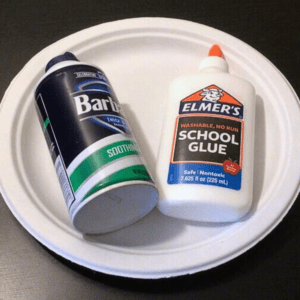 A can of Barbasol shaving cream and a bottle of Elmer's school glue sit on a white paper plate, placed on a dark surface to be used for kids crafts.