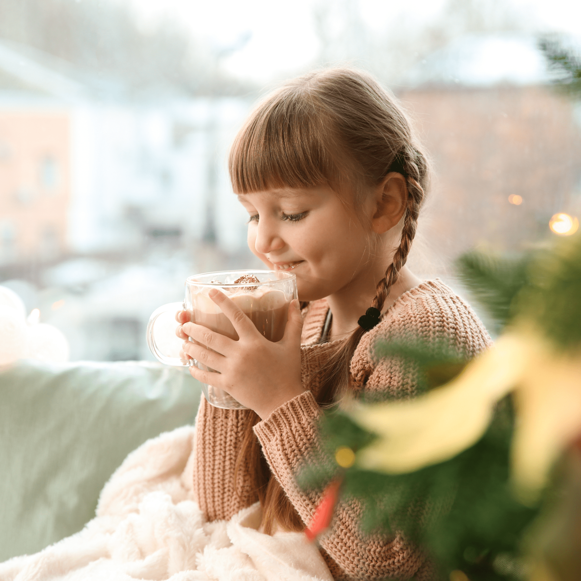 A young girl with braided hair smiles while holding a mug of hot chocolate, wrapped in a blanket by a window. A Christmas tree is in the foreground. It's winter story time!