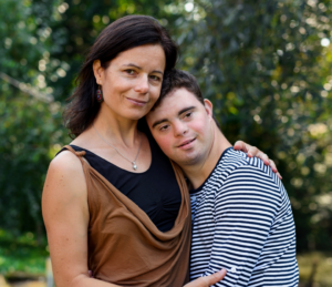 A woman and a young man with Down syndrome stand closely together in a sunlit park, smiling gently, surrounded by blurred green foliage. The mood is warm and loving.