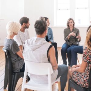 A group therapy session with eight people sitting in a circle on chairs in a bright room. One person speaks, others listen intently, fostering an open, supportive atmosphere.