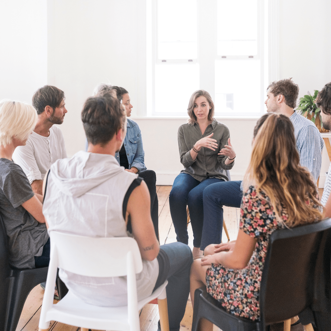 A support group sits in a circle in a cozy room with plants and soft lighting. A woman in the center appears emotional, while others offer comfort.