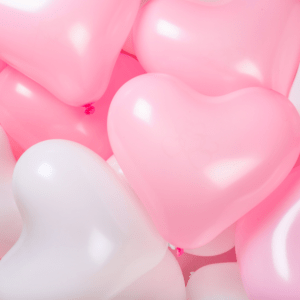 Heart-shaped balloons in soft pink and white fill the frame, creating a playful and romantic atmosphere. The shiny surface reflects light gently.