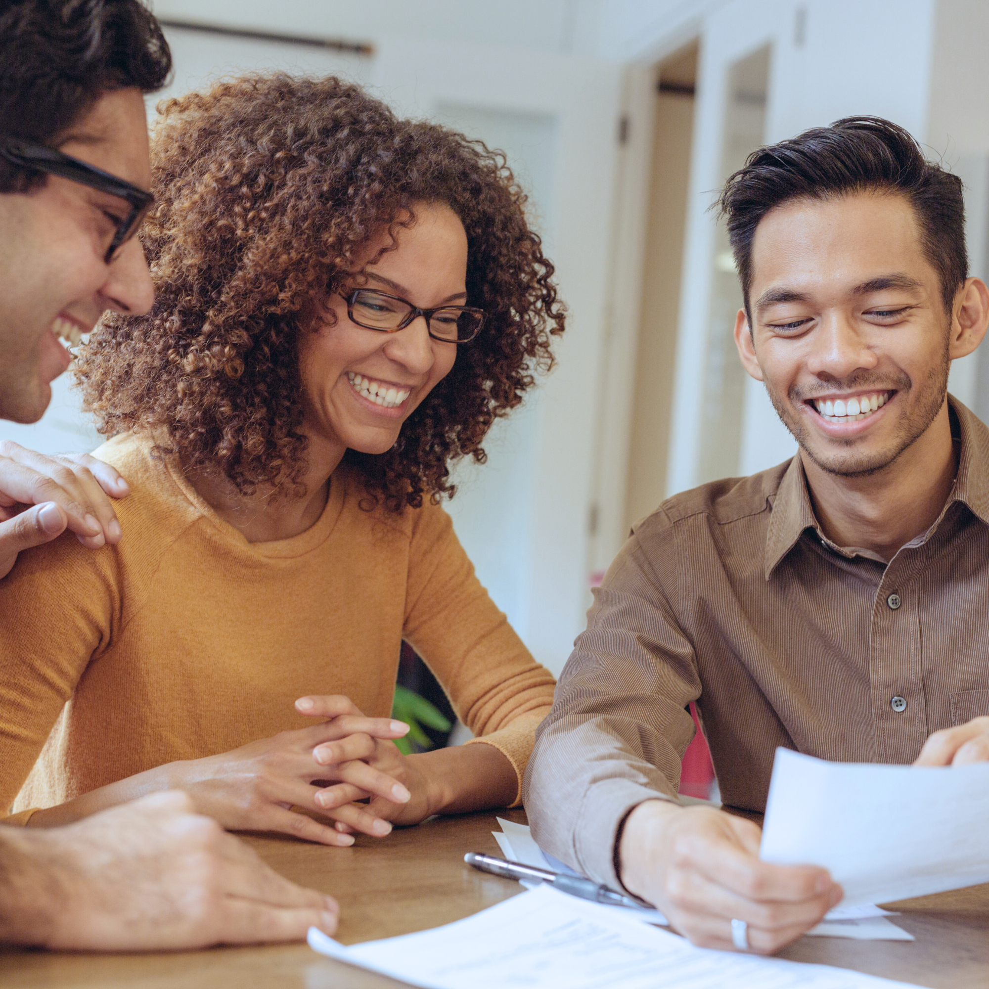 A diverse group of three individuals, teacher and parents, smiling and reviewing documents at a table, conveying a sense of teamwork and positive collaboration in a bright setting.