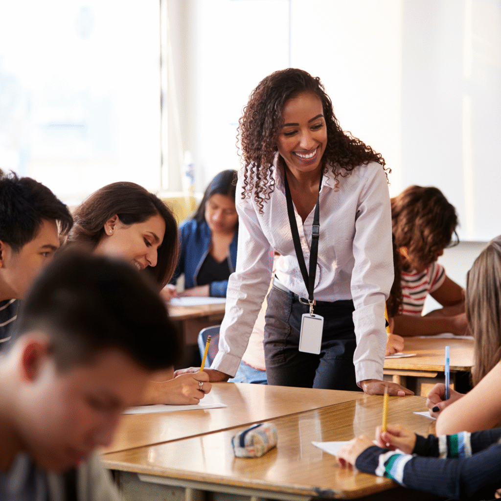 A smiling teacher leans over a desk, engaging with attentive students writing in a classroom. The atmosphere is positive and collaborative.