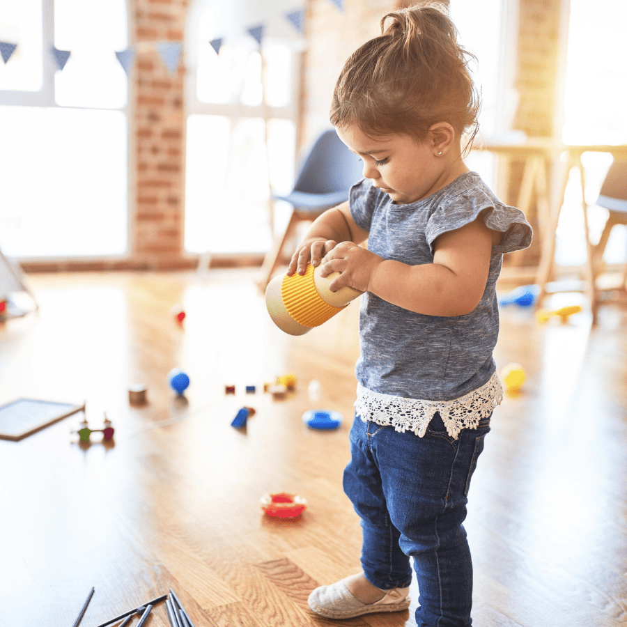 A toddler in a blue shirt and jeans plays with a yellow cup in a sunlit room. Toys are scattered on the wooden floor, conveying a playful, warm atmosphere.